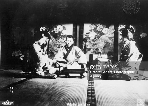 Two Japanese Geisha girls prepare tea for a nobleman in a scene from an unknown film.