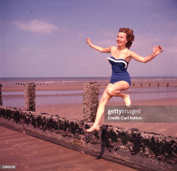 Marianne Brauns jumping a breakwater on the beach, wearing a structured blue swimsuit with white trim.