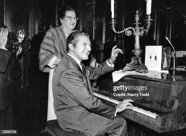 American pianist Liberace with his mother at a press reception at the Savoy Hotel, London. His trademark, a candelabra, is on the piano he is playing.