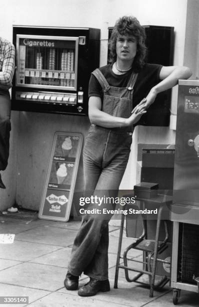 Model wearing dungarees, and a beaded necklace. He is standing near a cigarette vending machine.