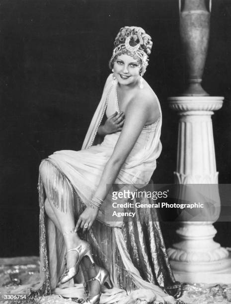 Thelma Todd the perky leading lady and heroine of many two-reel comedies is pictured sitting by a pillar and urn, wearing a very glamorous outfit...