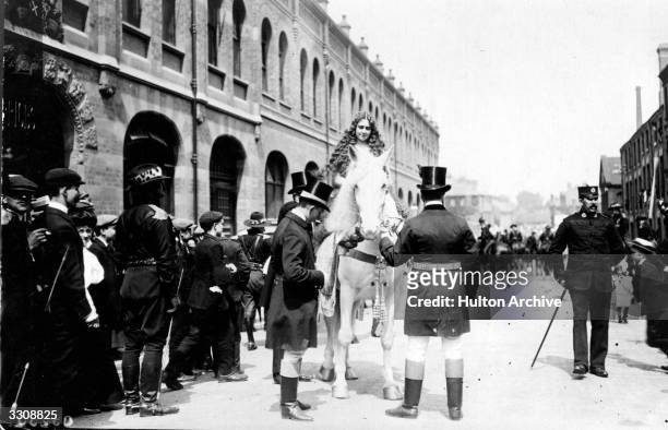 Lady Godiva mounts a fine white horse for a pageant in Coventry and a crowd of people look on.