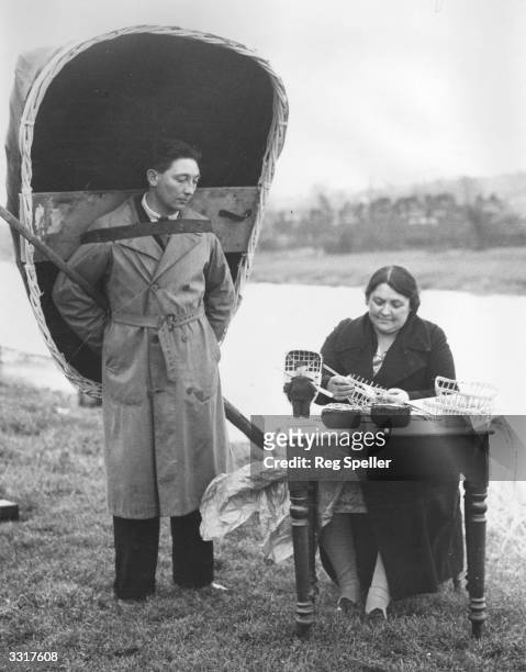 Mrs M Elias of Carmarthen, Wales, makes toy coracles, while her brother, holding a real coracle, models for her. She works on the bank of the river...