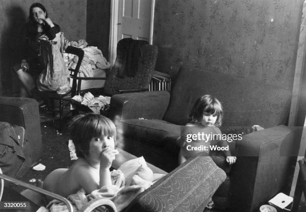 Couple of nude toddlers and their mother in their untidy sitting room.