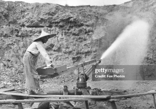 Worker at a tin mine in Malaya operating a water cannon.
