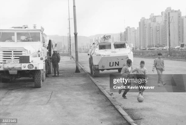 Children playing football beside UN vehicles parked at Sarajevo during civil war in Yugoslavia.