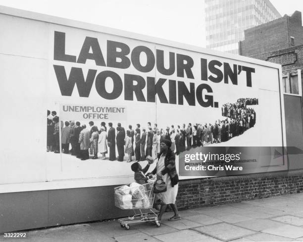 Woman with her shopping and little boy in a shopping trolley walk past a poster supporting the Conservative Party during the run up to the General...