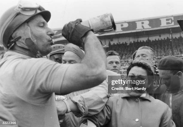 Argentinian racing driver Juan Manuel Fangio has a drink after winning the British Grand Prix at Silverstone. Watching him is his partner Andrea....
