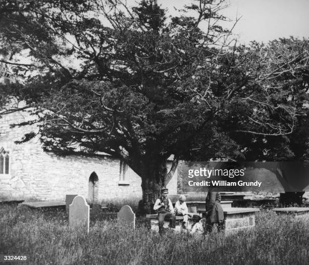 Yew tree gives shelter to a group sitting on the gravestones in an English churchyard. William Grundy's 'English Views'