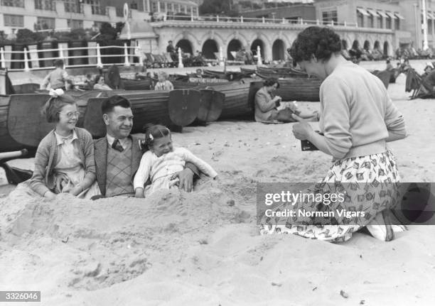 Fully clothed man and his two daughters pose buried in sand whilst his wife takes a photograph of them.