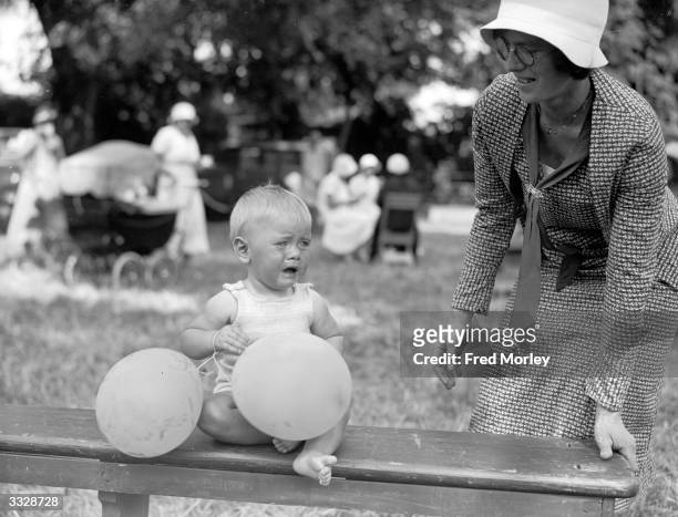 An unhappy baby holding balloons at a baby show, during a garden fete held at Cheam in Surrey, England, in aid of a local nursing association.