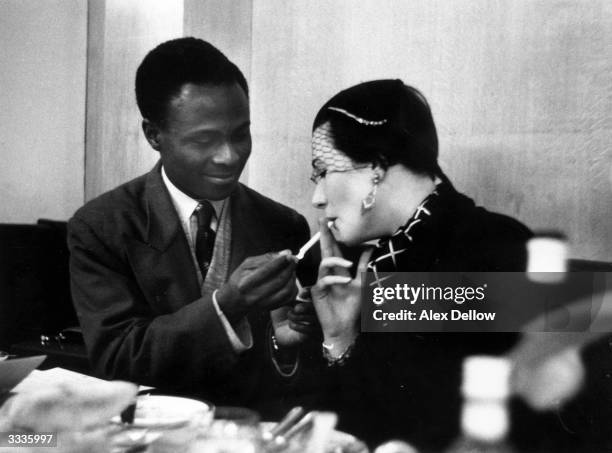 Nigerian student lights a cigarette for Patricia Farrier of South Africa at a London restaurant, February 1955. Farrier, who has left her native...