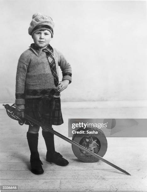 Little boy, Alex Mackintosh, dressed in a kilt and a knitted bonnet, beside a tin of Mackintosh's toffee, in a publicity shot for the brand. He is...