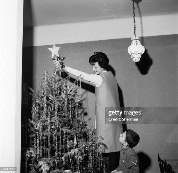 Woman puts a star on top of a Christmas tree, watched by her son.