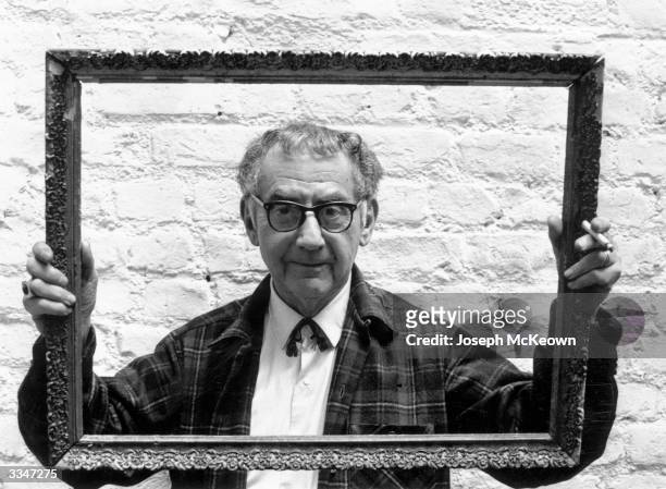 Painter, photographer and film-maker Man Ray peering through a picture frame which he is holding up as he stands against a whitewashed wall.