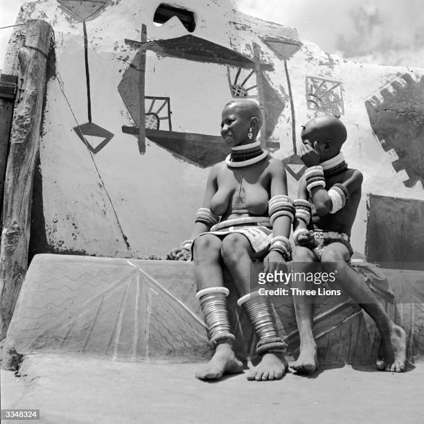 Two girls of the Ndebele tribe wearing brass bracelets around their ankles and sitting on a decorated wall in their village.