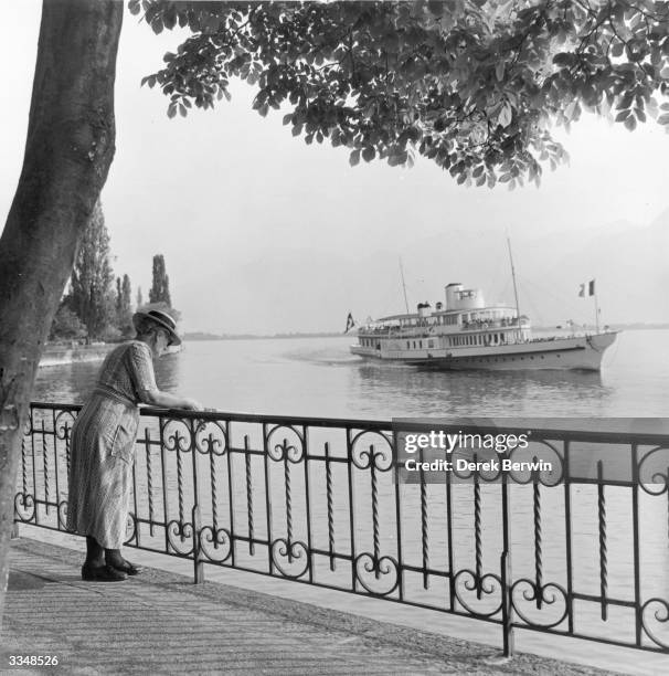 An elderly woman leaning on railings by the side of Lake Geneva at Montreux, where a steamer is passing by.