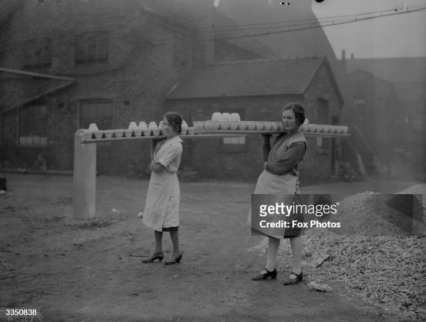 Two women balancing large trays of pots on their shoulders as they carry them across a yard at the Wedgwood pottery, Stoke On Trent, Staffordshire.
