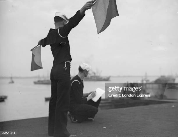 Sailor on shore signals to ship by semaphore, whilst another takes notes.