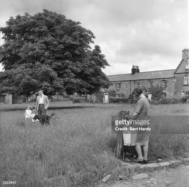 Woman filling a bucket with water at a tap on the village green at Wark, the old captial town of North Tyne, while a man and two little girls walk by...