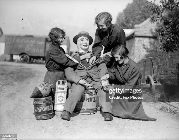 Three young women preparing a guy. They are sitting him on a keg labelled 'gun powder'.