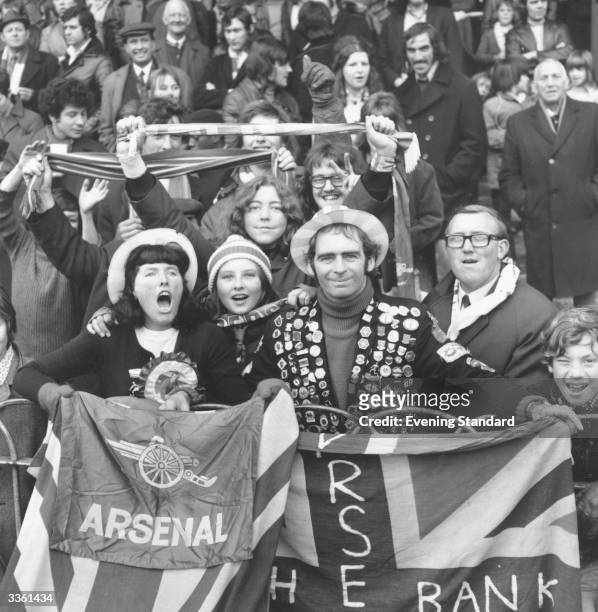 An Arsenal football club supporter holding a union jack and wearing a jacket festooned with badges with other fans during a match.