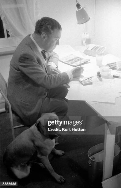 English couturier and court dressmaker Sir Norman Hartnell at work on designs in his salon at Bruton St, Mayfair, London, a bulldog by his side....