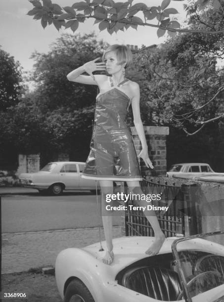 British model Twiggy wearing a transparent plastic halterneck dress, standing on the boot of a car.