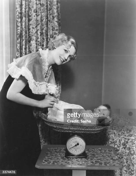 British character actress Enid Stamp-Taylor weighing her baby daughter Robin Anne in a set of scales at home at Maida Vale, London.