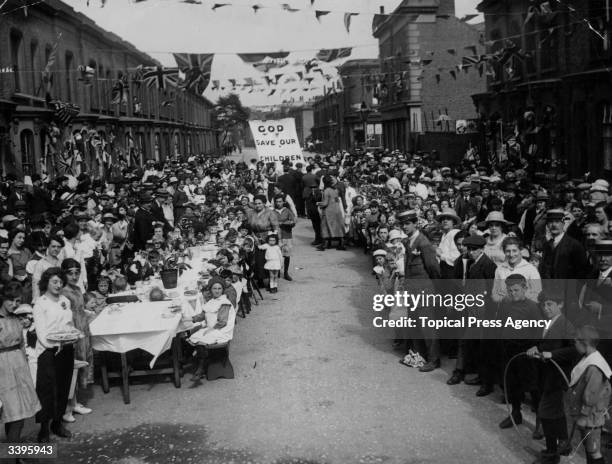 Crowds sitting down at long tables, at a street party to celebrate the end of the Great War.