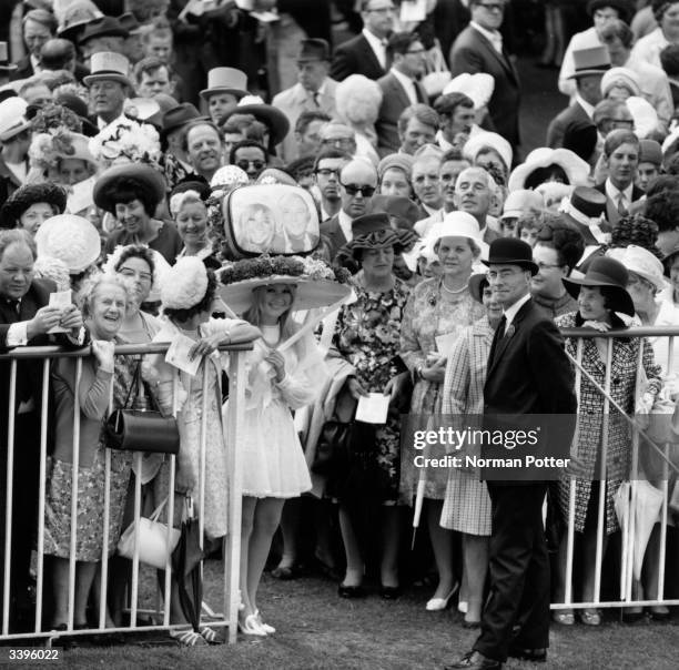 Crowds watching the Royal Procession at Ascot racecourse. One spectator is wearing an unusual hat.