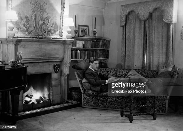 Couturier and court dressmaker Norman Hartnell at home by the fireside.