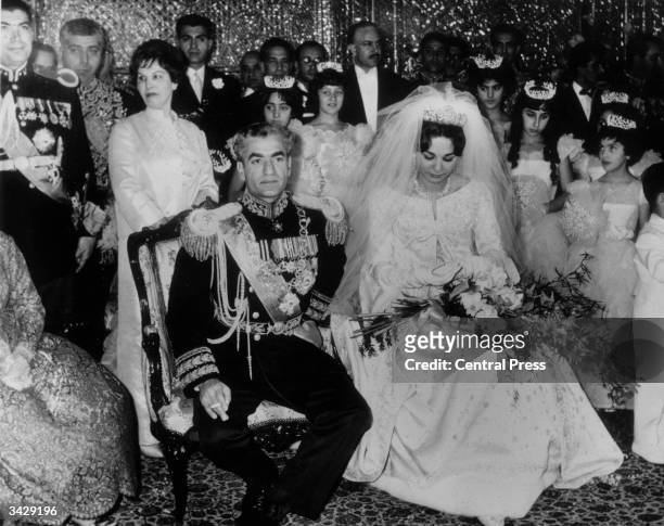 The Shah of Persia, cigarette in hand, wears the blue uniform of Commander-in-Chief as he sits beside his bride Farah Diba wearing a Dior dress of...