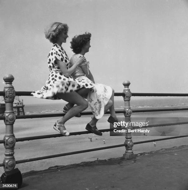 Two young women enjoy a summer day on Blackpool promenade as they sit on the railings with the wind billowing their skirts. They are former Tiller...