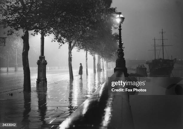 Man standing alone on a rain-drenched pavement on the River Thames Embankment, London.