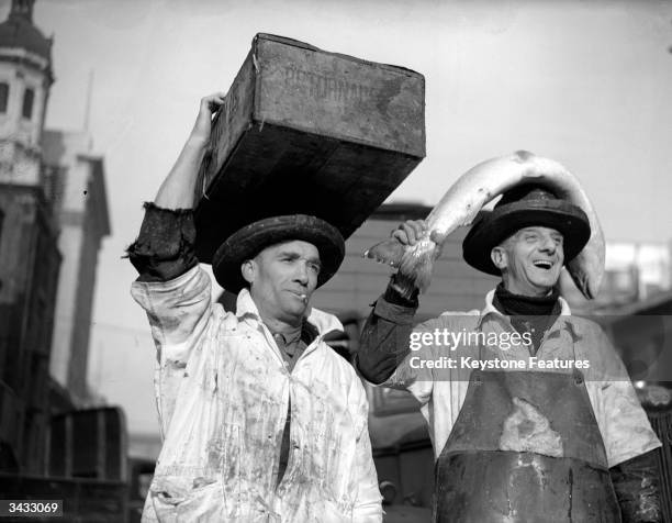 Two fish porters at Billingsgate fish market in London, one carrying a crate of fish and the other with a large salmon on his head.