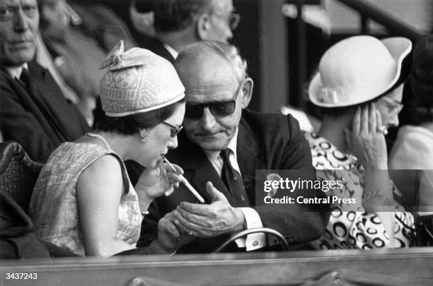 Wearing a matching dress and hat Princess Margaret , sister of Queen Elizabeth II, accepts a light for her cigarette from her companion in the royal...