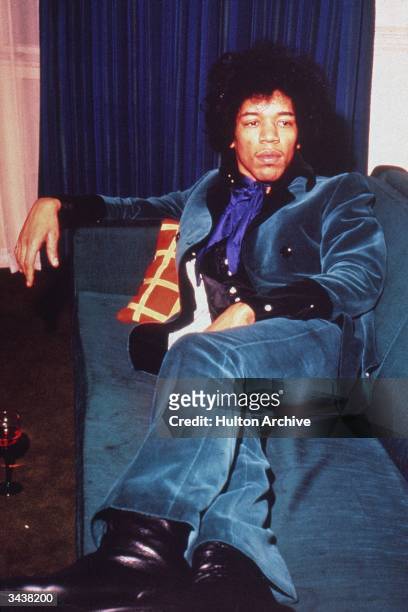 American rock musician Jimi Hendrix wears a teal velvet suit while reclining on a sofa.