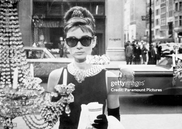 Belgian-born actor Audrey Hepburn , as Holly Golightly, holds a cup and a paper bag while looking into one of the window displays at Tiffany's in a...