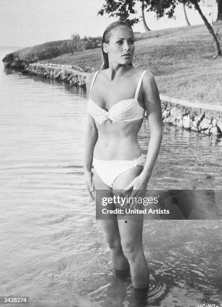 Swiss actor Ursula Andress wears a bikini while standing in water near the shore in a still from the James Bond film, 'Doctor No,' directed by...