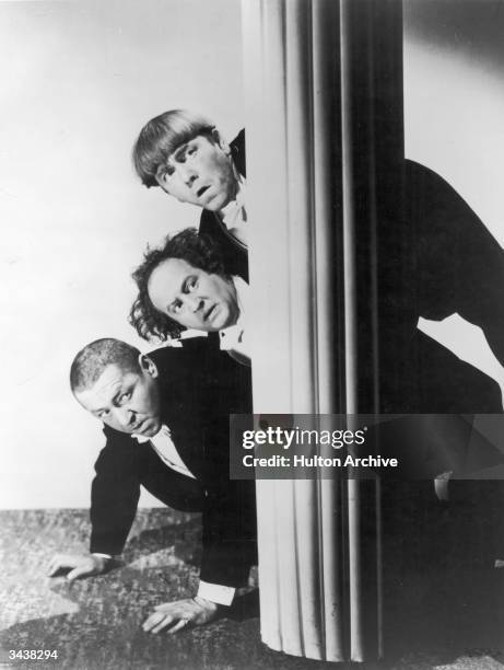 The Three Stooges peer around a column while hiding. Top to bottom: Moe Howard , Larry Fine and Curly Howard .