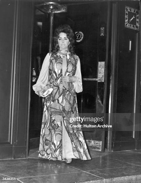 British actor Elizabeth Taylor, wearing a beaded maxidress, exits a revolving door.