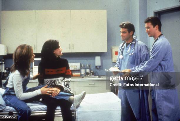 Jennifer Aniston and Courteney Cox sit in a hospital room, speaking to guest stars George Clooney and Noah Wyle from 'ER' in a still from the...