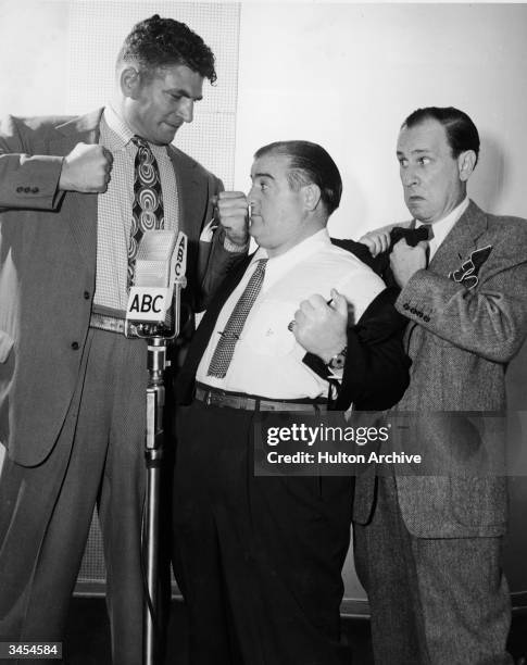 American comedy team Bud Abbott and Lou Costello cower as Lou Nova raises his fists at a microphone in a promotional still for a radio broadcast,...