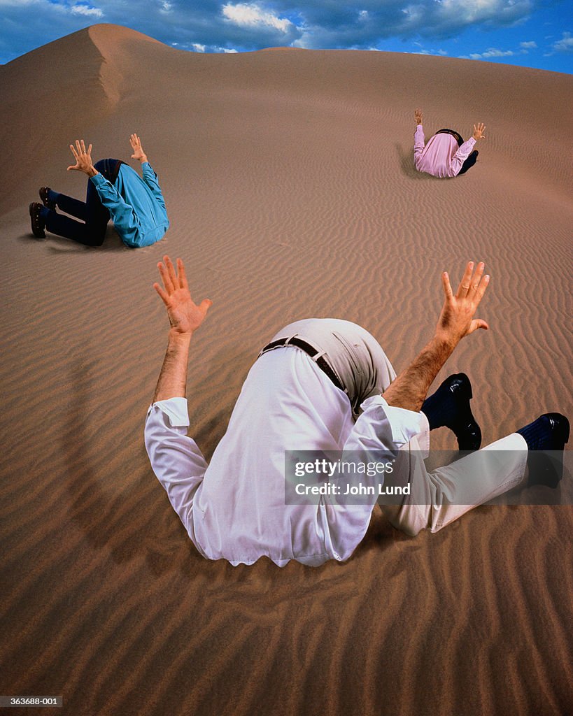 Three men with heads buried in sand dune (Digital Composite)