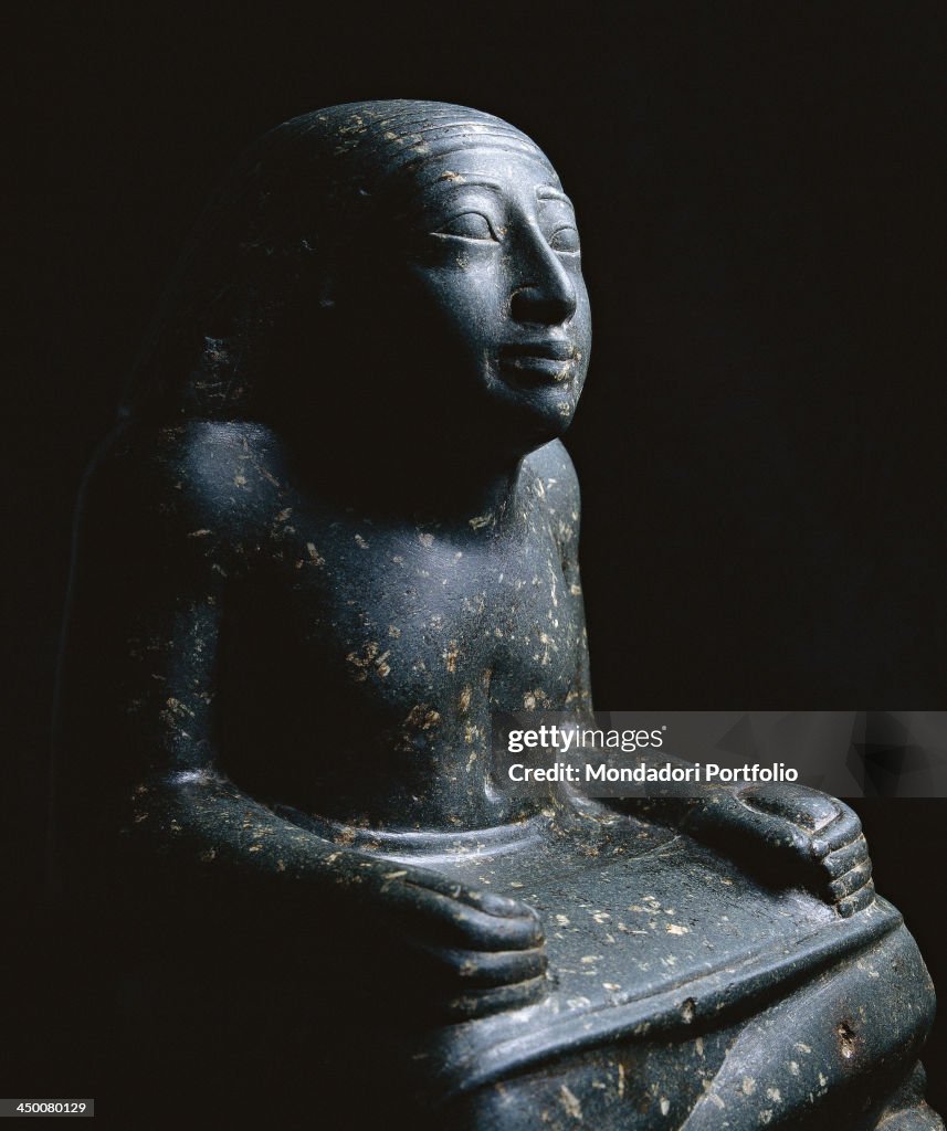 Private Collection. Detail. Three-fourths view of a male scribe with a wig on his head and a sheet of papyrus on his knees.