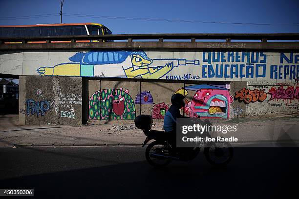Motorcycle rider passes a mural that shows the World Cup mascot shooting a rifle next to the words, "We want education not repression," as graffiti...