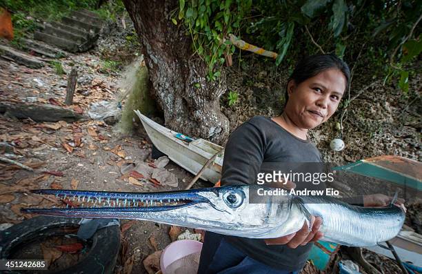 Women is holding one of the fish caught today by the local fishermen offering it for sale. The fish looks like a barracuda or needlefish with its...