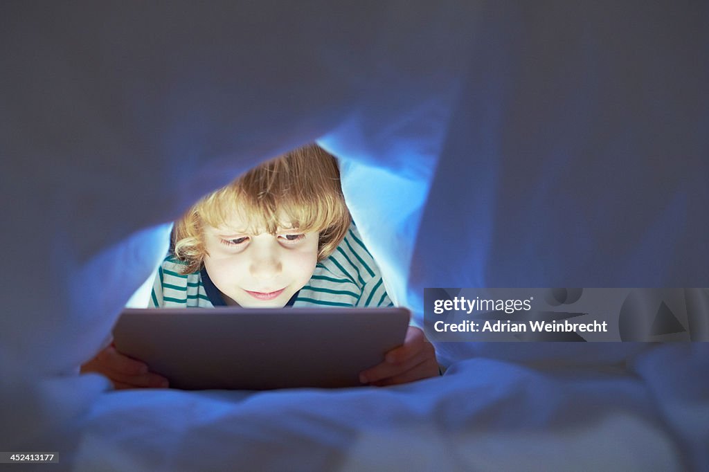 Boy underneath duvet using digital tablet