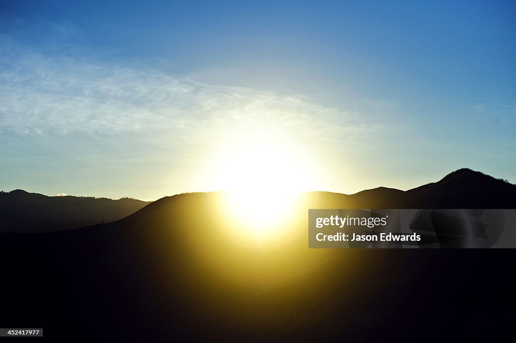 Dawn rises over a mountain ridge-line beneath a clear blue sky.